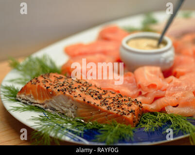 Gebratenes Lachsfilet auf einem Teller mit Dill angeordnet, Scheiben geräucherter Lachs und eine kleine China Schale mit Senfsauce Stockfoto