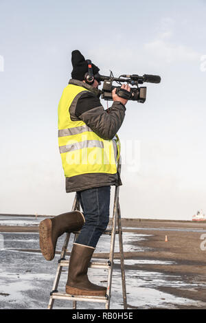 Kameramann mit gelber Weste auf steht auf einer Leiter am Strand Stockfoto