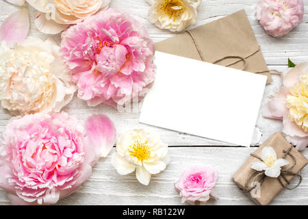 Leere weiße Grußkarte im Rahmen der Pfingstrose und Rosen Blumen und Umschlag mit Geschenkbox auf weißem Holz- Hintergrund. Flach. top anzeigen. Mock up Stockfoto