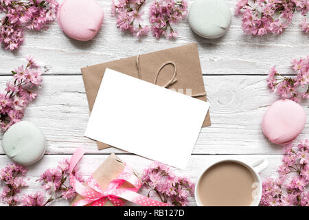 Leere Grußkarte oder Hochzeit Einladung im Rahmen der Pink Cherry Blüte mit Kaffeetasse und macarons über weiße Holztisch. Mock up. Stockfoto