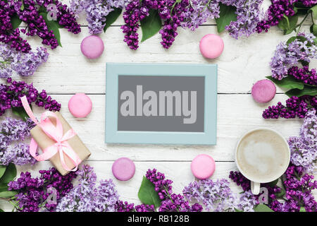 Leer Foto Karte im Rahmen aus lila Blüten mit Kaffeetasse, Geschenkbox und Makronen über weiße Holztisch. Mock up. Flach. top anzeigen. Mütter Stockfoto