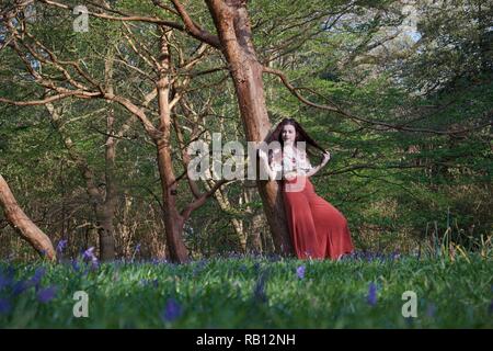 Eine modisch gekleidete junge Dame mit langen brünetten Haaren und roten Leuchtraketen lehnt sich gegen einen Baum und hält Ihr Haar. Die englische Waldland ist mit Teppichboden Stockfoto