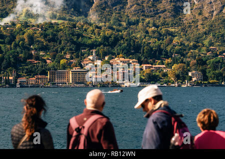 Cadenabbia, Italien - 7. Oktober 2017: Touristen beobachten, wie Boot fährt gegen Cadenabbia Stadt mit Gebäuden und Hotels, Küste der schönen Comer See, Es Stockfoto