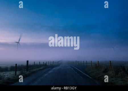 Ein Feldweg überschrift in die Ferne, als drei Windkraftanlagen steigen aus dem Nebel an einem nebligen Morgen. Stockfoto