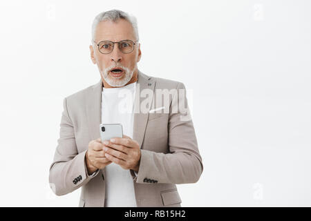 Taille - geschossen von schockiert älterer Mann in Anzug und Brille Keuchen öffnen den Mund von Sorgen und Überraschung holding Smartphone empfangen von schlechten Nachrichten suchen und Kamera über graue Wand betäubt Stockfoto