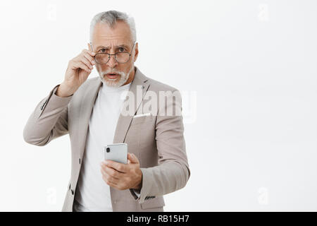 Portrait von überrascht und zögernd, alter Mann mit weißem Bart und Schnurrbart, die Bilder von der Kamera mit Handy verwechselt werden, wie Smartphone funktioniert über graue Wand, die versuchen, auf der Suche unsicher Stockfoto