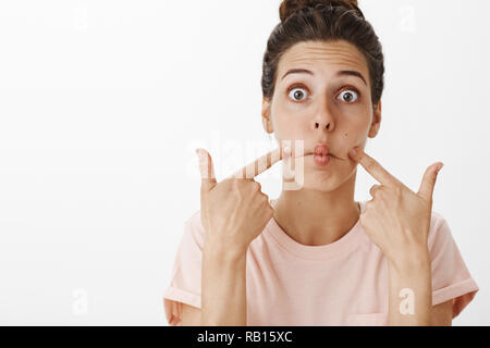 Taille-up Shot von überrascht cute woman als saugenden Lippen und Fisch Mund stoßend Wangen hebt Augenbrauen und popping Augen vor Erstaunen, Aprilscherze gegen grauen Hintergrund amüsiert sein Stockfoto