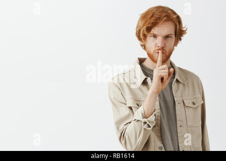 Shh lassen sie geheim. Portrait von geheimnisvollen gut aussehende junge Rothaarige kaukasischen Kerl in beige Jacke über t-shirt, psst Geste mit dem Zeigefinger auf den Mund schauen ernst Stockfoto