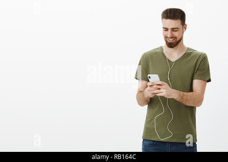 Studio shot der gut aussehende stilvolle bärtigen Kerl in Ohrhörer holding Smartphone und freudig lächelnd an mobilen Bildschirm Videos Ansehen in der Öffentlichkeit tragen Kopfhörer nicht Leute um stören Stockfoto