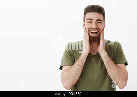 Wangen weh vom Lachen und Lächeln. Portrait von amüsiert glücklich upbeat attraktive Bärtigen männlichen Erwachsenen in Oliv t-shirt berühren Gesicht und grinste Spaß in super Stimmung auf weißem Hintergrund Stockfoto