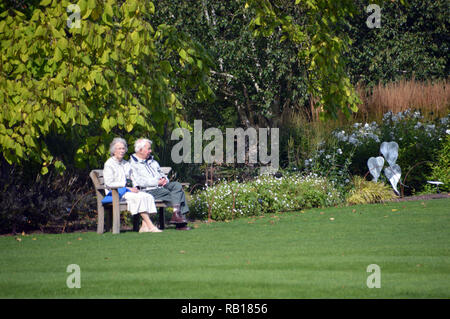 Reifes Paar sitzt auf einer Holzbank, auf einer Wiese im Sonnenschein an RHS Garden Harlow Carr, Harrogate, Yorkshire. England, UK. Stockfoto