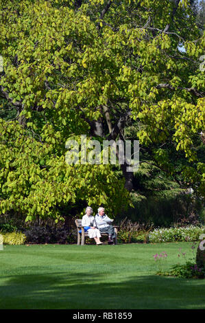 Reifes Paar sitzt auf einer Holzbank, auf einer Wiese im Sonnenschein an RHS Garden Harlow Carr, Harrogate, Yorkshire. England, UK. Stockfoto