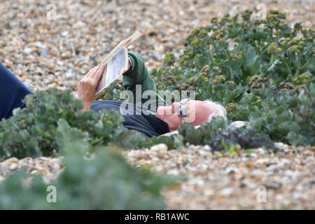 Ein Mann auf dem Rücken am Strand ein Buch lesen an einem kalten Tag im Frühling, in Großbritannien. Stockfoto