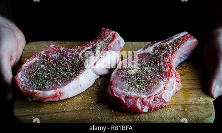 Hände halten einen Vorstand, an dem zwei rohes Schweinefleisch Steaks mit Gewürzen und bereit zu Kochen bestreut sind Stockfoto