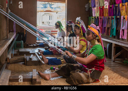 Vier Kayan (padaung) Frauen tragen Messing Hals Spulen und farbenfrohen traditionellen Kleid, Weberei Tuch mit traditionellen Webstühlen mit einer Pagode im Hinterg Stockfoto