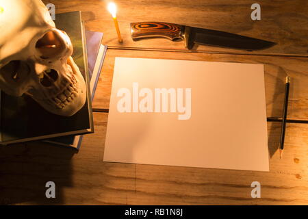 Menschlicher Schädel mit Kerzenlicht und weißes Papier mit Bleistift und Messer auf hölzernen Tisch, Dekoration für Halloween Theme mit kopieren. Stockfoto