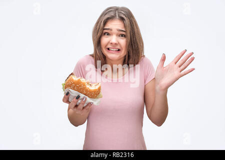 Erschrocken und frustrierte junge Frau, gebissen Burger in der Hand. Sie zeigt Palm. Auf grauem Hintergrund. Stockfoto