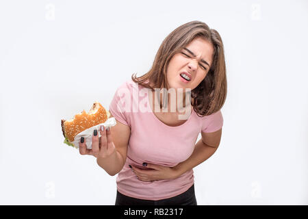 Junge Frau leiden unter Schmerzen im Magen. Sie gebissen Burger. Eine andere Hand ist auf Bauchpartie. Auf weissem Hintergrund. Stockfoto