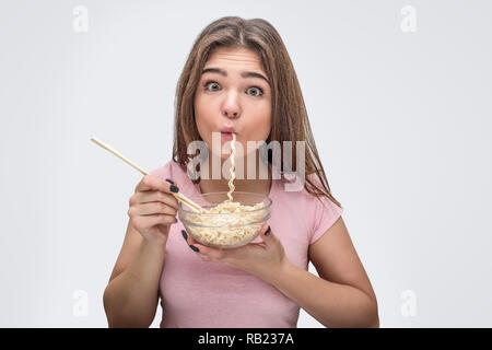 Lustige junge Frau hat einige Nudeln in den Mund. Sie fressen es gerne. Modell halten Schüssel mit Nudeln in der Hand. Auf weissem Hintergrund. Stockfoto