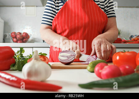 Ältere Frau kochen zu Hause in der Küche, schneiden Zwiebeln auf ein Holzbrett. Gesunde frische Lebensmittel. Stockfoto