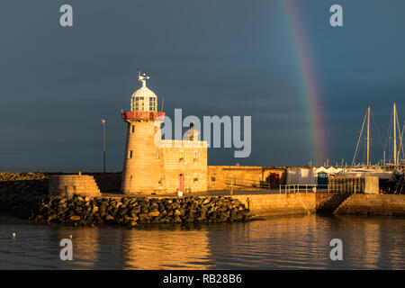 Der Leuchtturm am Hafen von Howth, County Dublin gebadet im Sonnenlicht unter einem Regenbogen vor einem stürmischen Himmel, mit seinem Spiegelbild im Wasser. Stockfoto