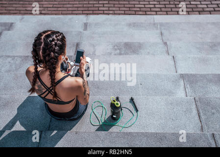 Mädchen mit Tätowierungen in Sportkleidung, Ansicht von oben, von hinten. Er hält ein Smartphone in seinen Händen, schreibt eine Nachricht an Internet. Sport und gesunde Lebensweise. Freier Platz für Text. Stockfoto