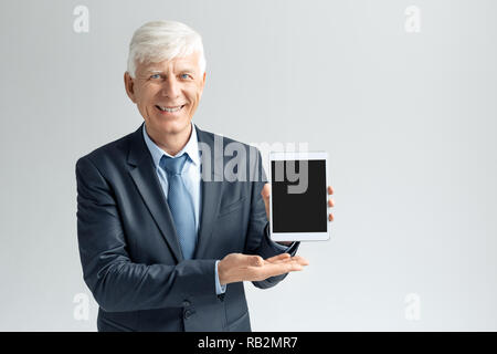 Senior Business mann Studio stehen isoliert auf graue Wand Holding digital Tablet angezeigt Bildschirm schaut die Kamera lächelt freundlich Stockfoto