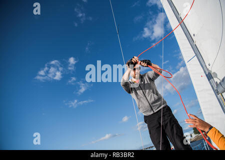 Gut aussehender bärtiger Matrose arbeiten mit Seilen an Deck einer Yacht gegen den klaren, blauen Himmel und Meer Stockfoto