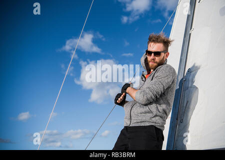 Gut aussehender bärtiger Matrose arbeiten mit Seilen an Deck einer Yacht gegen den klaren, blauen Himmel und Meer Stockfoto