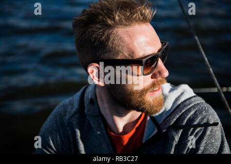Close up Portrait bärtiger Mann Modell mit Sonnenbrille und Kapuze schaut weg, den Sonnenuntergang im Meer. Stockfoto