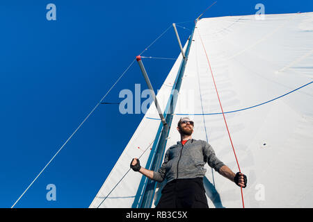 Schöne und selbstbewusste junge Mann steht auf Yachtcharter board und im Meer durch die Brille schauen. Er liegt am Rande der Yacht. Guy Holding segeln Seil in der Hand. Junge Mann ist glücklich und friedlich. Stockfoto