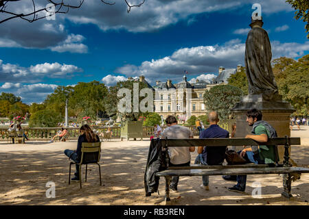 Ein Mädchen sitzt auf einem grünen Stuhl prüfen Ihr Telefon whist in der Nähe drei Männer sitzen auf einer Bank an einem Sommertag im Jardin du Luxembourg, Paris, Stockfoto