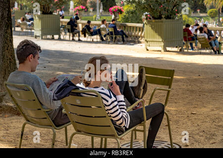 Eine junge Frau, die auf einem grünen Stuhl sitzt und eine Zigarette raucht, neben einem Mann, der eine Zeitung liest, an einem Sommertag im Jardin du Luxembourg, Paris, Frankreich Stockfoto