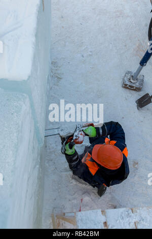 Workman Assembler in einem Schutzhelm mit einer Kettensäge in der Hand schneidet den Eisblock Stockfoto