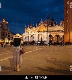 Touristen fotografieren den Markusdom bei Nacht auf der Piazza San Marco, Venedig, Italien, mit beleuchteter Architektur und tiefblauem Abendhimmel. Stockfoto