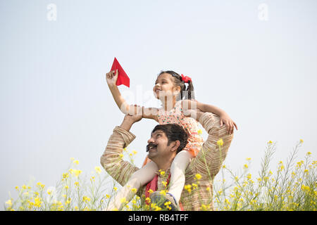 Ländliche indische Mädchen sitzt auf der Schulter des Vaters und dem Spielen mit Spielzeug Papierflieger im Feld Stockfoto