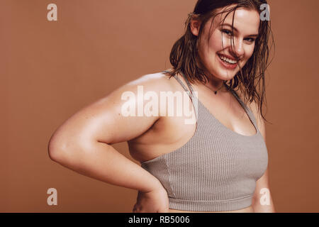 Plus Size weibliche Modell im Sport-BH mit Kamera und lächelnd. Positive übergroße jungen kaukasischen Frau gegen braunen Hintergrund. Stockfoto