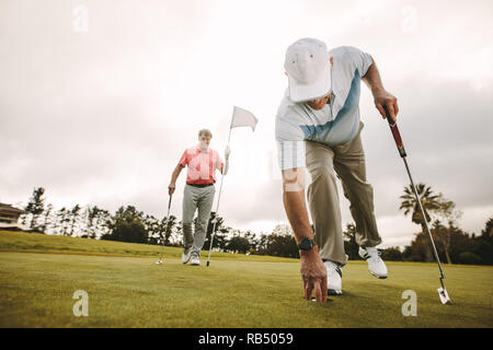 Ältere Golf player Kommissionierung Golf Ball aus der Bohrung mit einem zweiten Spieler im Hintergrund halten die Flagge. Ältere Golf Spieler ein Spiel spielen auf dem Golfplatz Stockfoto