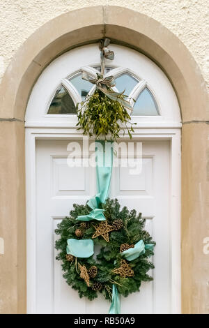 Weihnachten kiefer Kranz mit Tannenzapfen und gold Sterne, auf einer weißen Tür von Green Ribbon aufgehängt. Oben ist es große Haufen von Mistel Stockfoto