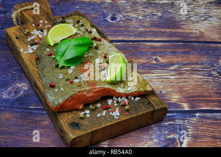 Raw Regenbogenforelle steak bestreut mit Gewürzen, Pfeffer, Salz mit Stücken von Kalk und Basilikum auf einem alten Holz- Board. Flache Ansicht von oben in der vinta Stockfoto