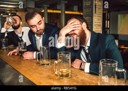 Junge bärtige Mann vor und Schlafen sitzen. Er ist betrunken. Zweite junge Mann schauen ihn an. Dritte Bier trinken. Stockfoto