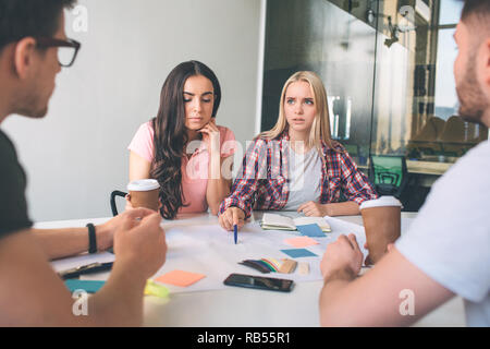 Ernst und besorgt, junge blonde Frau Blick auf Männer. Sie sitzen mit Ihnen und Ihrer Freundin an einen Tisch. Blonde Modell Punkt auf Stück Papier. Kerl auf Sie. Stockfoto