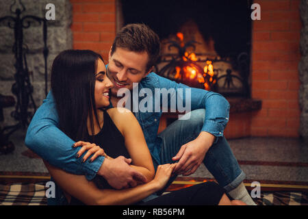 Fröhliche junge Frau zusammen mit der man auf dem Boden sitzen. Er ihre Umarmung. Sie sitzen gerne in der Nähe der Feuerstelle. Stockfoto