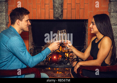 Fröhliches Bild der junge Mann und die Frau einander sitzen auf Stühlen. Sie jubeln mit Champagner Gläser. Paar sitzen am Kamin. Stockfoto