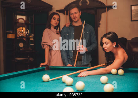Attraktive junge asiatische Frau spielen Billard Spiel. Sie sieht Noce und konzentriert. Ihre Freunde stehen. Sie sehen Ihr Spiel Stockfoto