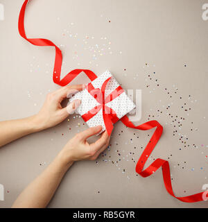 Geschenkbox mit roter Schleife in der Frau die Hand und Konfetti auf grauem Hintergrund. Valentinstag Muttertag, Verkauf, Geburtstag Konzept Stockfoto