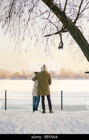 Thema Liebe und aktive Erholung auf rustikalen Charakter. Junges Paar kaukasischen Kerl und ein Mädchen stehen wieder im vollen Wachstum Hand in Hand im Winter auf Schnee pier overlo Stockfoto