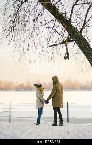 Thema Liebe und aktive Erholung auf rustikalen Charakter. Junges Paar kaukasischen Kerl und ein Mädchen stehen wieder im vollen Wachstum Hand in Hand im Winter auf Schnee pier overlo Stockfoto