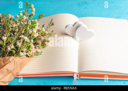 Kleine weiße Blumen Blumenstrauß auf ein leeres Notebook und zwei hölzerne Herz, auf blauem Hintergrund. Valentinstag Geschenke. Glückliche Momente. Jubiläum. Stockfoto