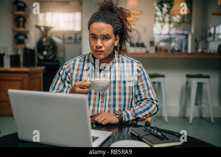 Portrait einer jungen schönen gemischten Rennen Kerl mit gebunden Afro, Halten einer Tasse Kaffee hinter seinem Laptop ausschalten Stockfoto